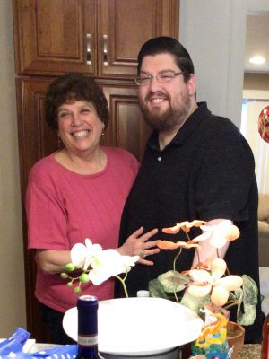 A woman in a pink shirt and a man in a black shirt stand together indoors, smiling beside a counter with flowers and a bottle in the foreground.