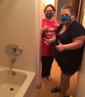 Two women wearing face masks stand in a hallway next to a bathroom, both giving thumbs up toward the camera.