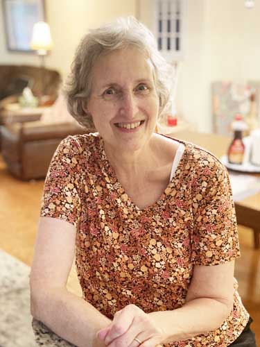 An older woman with short gray hair and a floral shirt is smiling while seated in a well-lit living room.