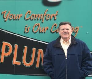 A man in a navy jacket stands and smiles in front of a green truck with the partially visible word "PLUMB" and the phrase "Your Comfort is Our Calling" on it.