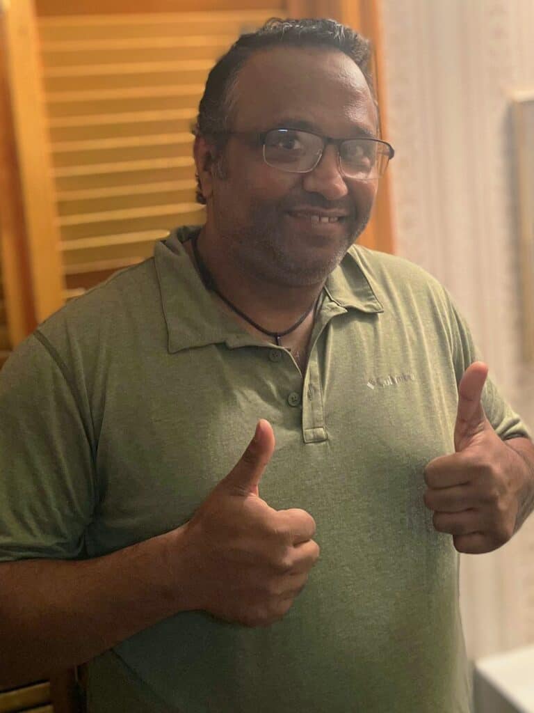A man wearing glasses and a green collared shirt gives two thumbs up while smiling at the camera indoors.