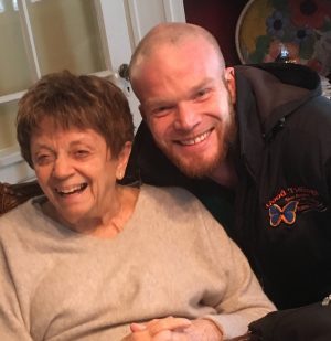 An elderly woman and a younger man smile as they pose together indoors. The woman is seated, and the man is leaning in beside her.