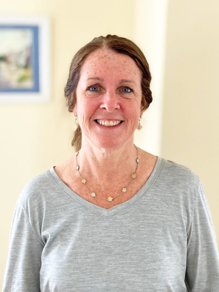 A woman with light skin, brown hair pulled back, and blue eyes smiles at the camera. She is wearing a light gray shirt and a gold flower necklace, standing indoors with a painting in the background.