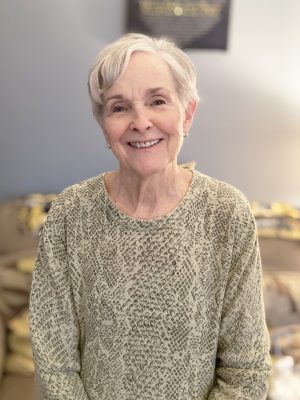 An older woman with short gray hair smiles at the camera, wearing a light patterned sweater, standing indoors in front of a sofa.