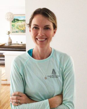 Woman wearing a light blue long-sleeve shirt with a logo stands indoors, smiling with arms crossed. A painting and table are visible in the background.