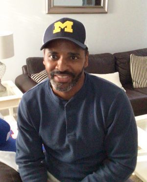 A man with a beard wearing a navy blue shirt and a cap with a yellow "M" sits on a couch in a living room.