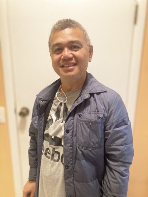 A man with short gray hair is smiling while wearing a blue jacket over a gray Reebok t-shirt, standing indoors in front of a white door.
