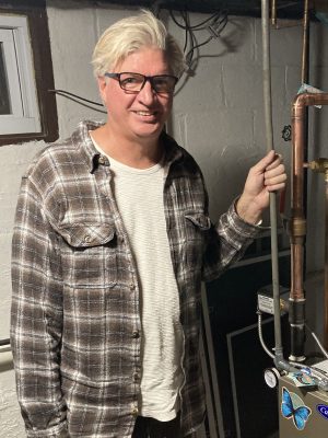 A man with light hair and glasses stands indoors near pipes and equipment, holding onto a metal pipe. He is wearing a plaid shirt over a striped t-shirt.