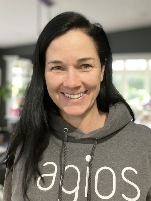 A woman with long black hair smiles at the camera, wearing a gray hoodie with white text on the front. Indoors, with blurred background.