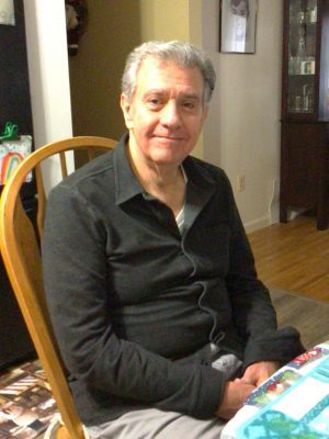 An older man with gray hair sits on a wooden chair at a table in a home setting, wearing a dark shirt and light pants, looking at the camera with a neutral expression.