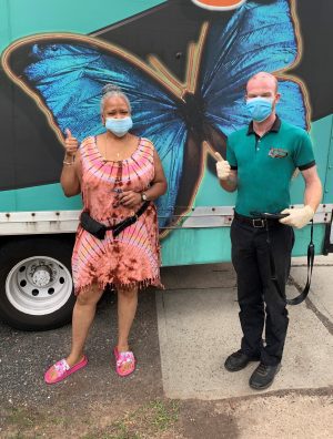 Two people wearing masks and gloves stand in front of a truck with a large blue butterfly graphic, both giving a thumbs-up gesture.