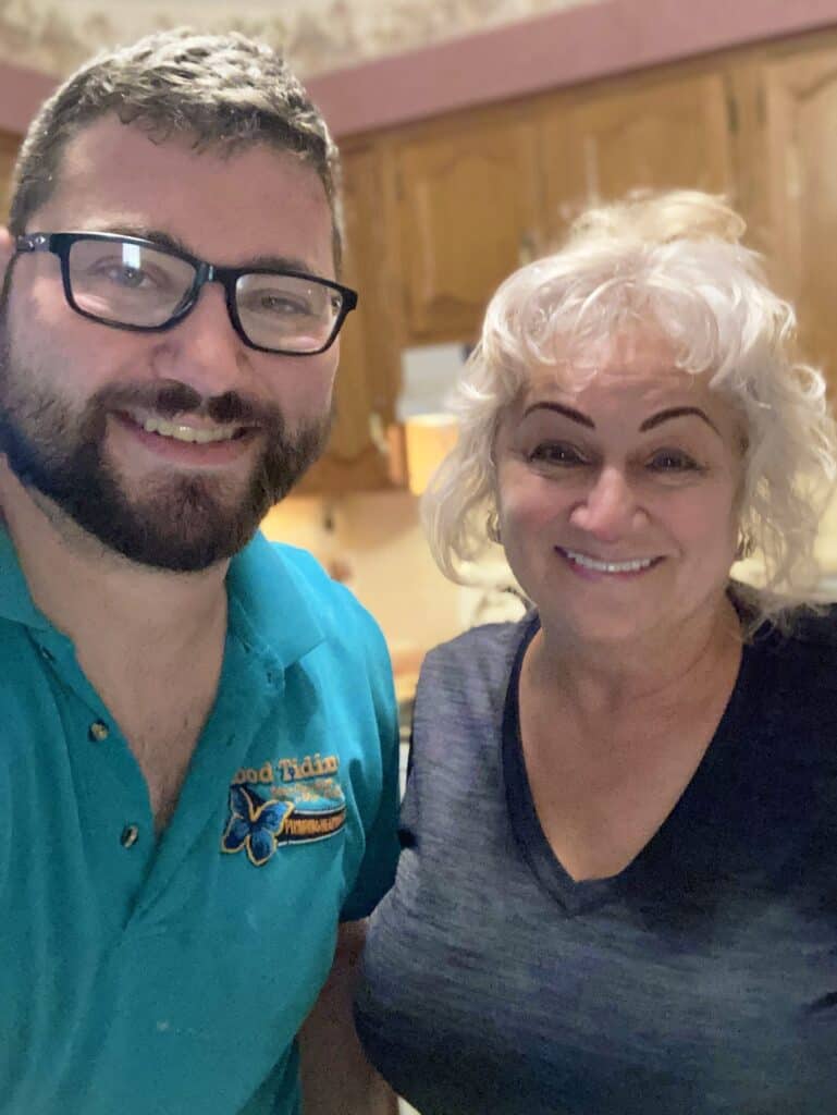 A man with glasses and a beard stands next to a smiling older woman in a kitchen with wooden cabinets.
