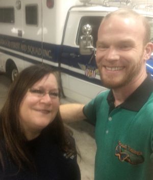 Two people are smiling for a selfie in front of an emergency medical vehicle inside a garage.