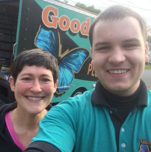 Two people smiling for a selfie in front of a truck with a large blue butterfly and the word "Good" visible on the side.