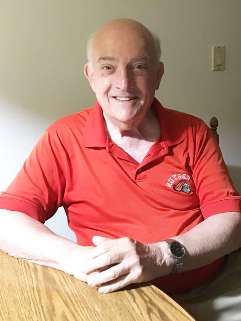 An older man wearing a red Rutgers polo shirt sits at a wooden table, smiling at the camera in a well-lit room.