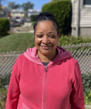 A woman wearing a pink hoodie stands outside in front of a chain-link fence and greenery, smiling at the camera.