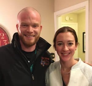 A man and a woman stand side by side indoors, both smiling at the camera.