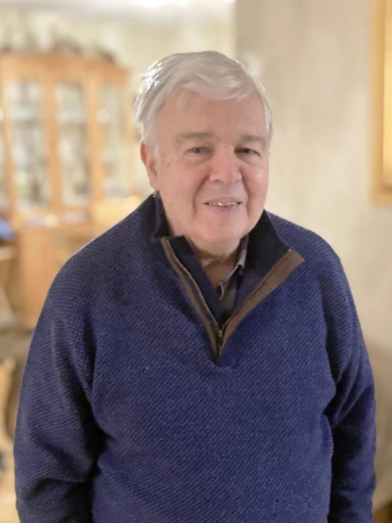 An older man with white hair wears a blue pullover and stands indoors in a softly lit room with a blurred background.
