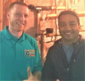 Two men standing indoors, one wearing a teal polo shirt with a logo and the other in a dark jacket, both smiling at the camera with pipes and wires in the background.