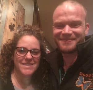 Two people posing together indoors, one wearing glasses and the other wearing a jacket with embroidered text and a dog graphic.