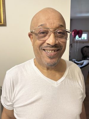 Smiling man with glasses and a white t-shirt stands indoors; red heart-shaped balloons are visible in the background.