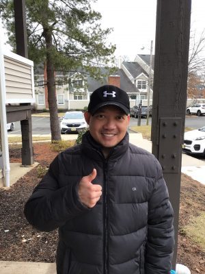 A man wearing a black cap and puffy jacket stands outside, smiling and giving a thumbs up. Houses, cars, and trees are visible in the background.