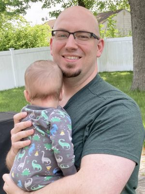 A smiling man wearing glasses holds a baby dressed in a dinosaur-patterned outfit outdoors near a white fence and trees.