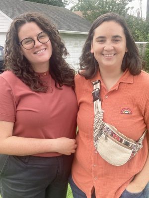 Two people stand outdoors in front of a garage, both smiling at the camera. One wears glasses and a red shirt, the other wears an orange shirt and a beige crossbody bag.
