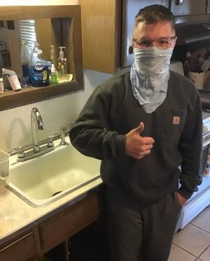 Person wearing glasses and a face covering gives a thumbs up while standing next to a kitchen sink in a home setting.