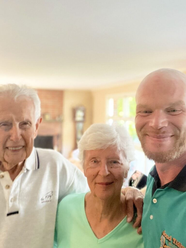 Three people stand close together indoors, smiling at the camera. An older man and woman are on the left, and a younger man is on the right. The background shows a bright, homey living room.