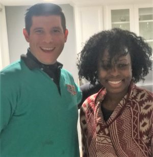 Two people standing indoors, smiling at the camera; one person wears a teal shirt and the other wears a patterned sweater.
