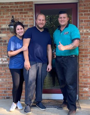 Three adults stand in front of a brick house entrance; two are casually dressed and smiling, while the third wears a teal shirt and gives a thumbs up.