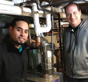 Two men standing next to a furnace unit in a basement with various pipes and insulation visible in the background.