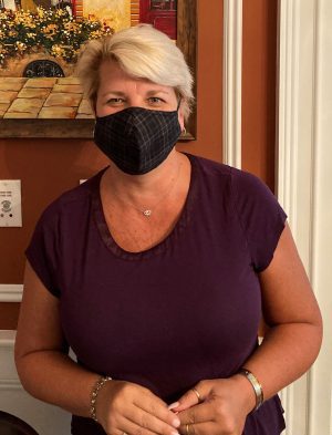 A woman with short blonde hair wearing a black face mask and a purple shirt stands indoors in front of a reddish-brown wall.