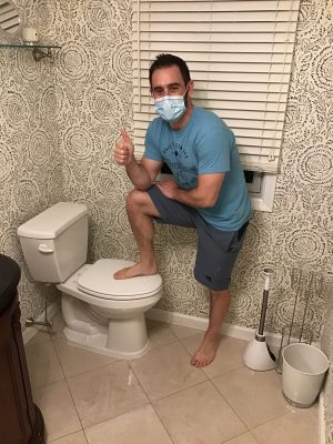 A man wearing a mask and casual clothes stands in a bathroom with one foot on a closed toilet seat, giving a thumbs-up gesture.