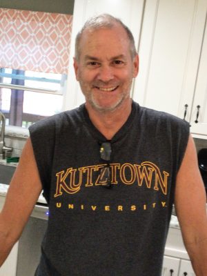 A man stands in a kitchen wearing a sleeveless Kutztown University shirt and smiling at the camera.