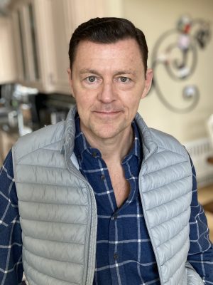 Man with short dark hair wearing a blue plaid shirt and light gray quilted vest stands indoors, facing the camera. Kitchen cabinets and wall art are visible in the background.
