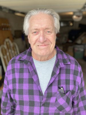 An older man with gray hair, wearing a purple and black plaid shirt over a gray top, stands in a garage and smiles at the camera.