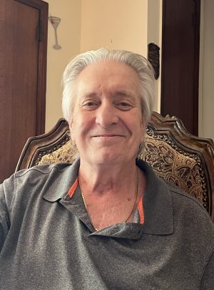 An older man with white hair and a gray collared shirt sits on a carved wooden chair, smiling at the camera indoors.