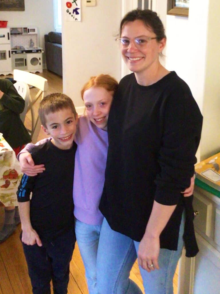Three people stand indoors, smiling at the camera. An adult with glasses stands on the right, while two children stand close together on the left, one with an arm around the other.