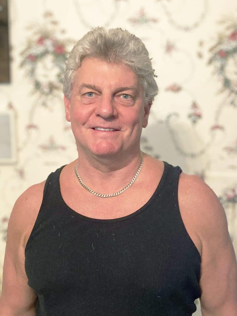 A man with gray hair wearing a black tank top and a silver chain necklace stands in front of a floral-patterned wallpaper.