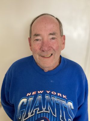 An older man with short hair smiles at the camera while wearing a blue "New York Giants" sweatshirt against a plain light background.