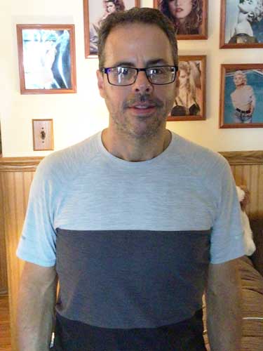 A man with glasses and short hair stands indoors in front of a wall with framed photos. He is wearing a light and dark color-blocked shirt.