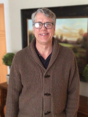 A man with gray hair and glasses wearing a brown knitted cardigan stands indoors, smiling at the camera, with a painting and plants in the background.