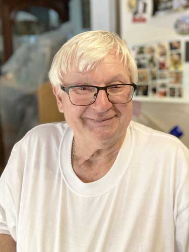 An older man with glasses and white hair smiles at the camera, wearing a white T-shirt. There are photos and items on the wall in the background.
