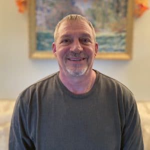 A middle-aged man with short gray hair and a goatee is smiling, wearing a gray shirt, seated indoors with a painting on the wall behind him.