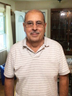 A middle-aged man wearing glasses and a light-striped polo shirt stands indoors near a window and a glass cabinet.