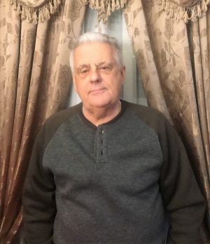 An older man with gray hair wearing a dark henley shirt stands in front of patterned curtains.