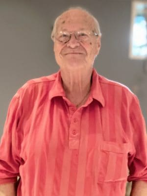 An older man with glasses is wearing a red striped shirt and smiling while standing indoors in front of a gray wall.