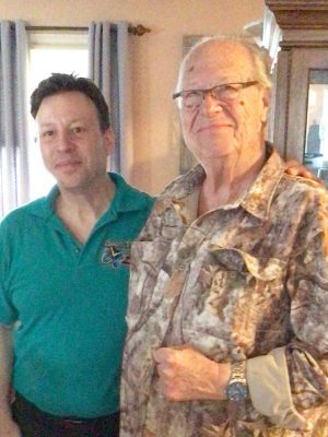 Two men standing indoors next to each other, one wearing a green polo shirt and the other wearing a camouflage shirt and glasses, both looking at the camera.